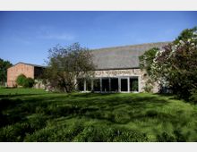 Großes Gebäude mit einer Fassade aus Naturstein und Metallfenstern, flankiert von Bäumen und blühenden Sträuchern in einer grünen Wiesenlandschaft; links im Hintergrund ein weiteres Gebäude aus rotem Backstein.