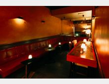 Schmaler Lounge- oder Barbereich mit langen roten Leder-Sitzbänken entlang der Wand, kleinen runden und eckigen Tischen sowie gedämpften Tischlampen. Warme, orangefarbene Wände und ein langer Tresen mit marmorierter Oberfläche prägen den Innenraum.