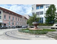 Ein kleiner Platz mit einem Brunnen in der Mitte, umgeben von einer verzierten Metallumzäunung. Im Hintergrund stehen ein rosa Gebäude mit Satteldach und ein moderneres Gebäude mit weißen Fassaden und großen Fenstern, daneben Bäume und parkende Autos.