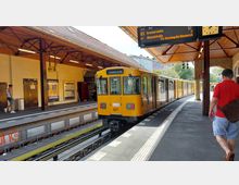 Gelber Berliner U-Bahn-Zug der Linie U3 mit Ziel "Krumme Lanke" steht an einem historischen überdachten Bahnhof mit Holzdecke und Metallpfeilern. Im Hintergrund ist eine digitale Anzeigetafel, eine Uhr, sowie städtische Gebäude und Grünflächen sichtbar.