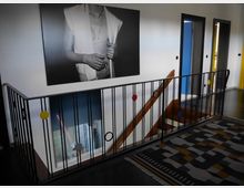 Treppenhaus mit einem schwarzen, modernen Geländer, das bunte Farbakzente aufweist. Im Hintergrund befinden sich drei Türen in verschiedenen Farben (blau und gelb) sowie eine großformatige Schwarz-Weiß-Fotografie an der Wand. Ein gemusterter Teppich bedeckt den Boden.