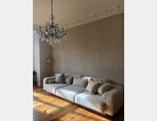 Beige Wohnzimmer mit einer großen, gepolsterten Couch, dekoriert mit Kissen in verschiedenen Farben. Im Raum gibt es einen Kronleuchter, eine verzierte Stuckdecke, Holzboden sowie eine Stehlampe und Pflanzen in der Ecke.