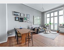 Helles Wohnzimmer mit Holzboden, grauer Wand und großen Fenstern, die viel Tageslicht hereinlassen. Ein Sofa mit Kissen, ein runder Couchtisch, ein Esstisch mit Stühlen sowie eine Wand mit gerahmten Bildern und Zimmerpflanzen bestimmen die Einrichtung.