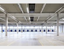 Große, leere Lagerhalle mit Betonboden, nummerierten blauen Toren an der Rückwand und einer hohen Decke mit sichtbaren Belüftungsschächten und Leuchtstoffröhren. Reihen von Betonsäulen stützen die Konstruktion.