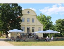 Gelbes, zweistöckiges Gebäude mit halbrundem Giebeldach, großen Fenstern und einer zentralen Treppe vor dem Eingang. Im Vordergrund stehen Tische und Bierbänke unter weißen Sonnenschirmen auf einem grasbewachsenen Platz, umgeben von Bäumen.