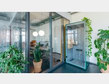 Moderner Büroraum mit großen Glaswänden, die einen Besprechungsraum mit Tisch und Stühlen abgrenzen. Im Vordergrund stehen mehrere Topfpflanzen sowie eine kleine schallisolierte Kabine mit Hocker und Schreibtisch; die Decke zeigt sichtbare Rohrleitungen.