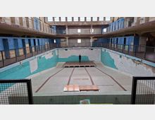 Ehemaliges Hallenbad mit leerem Becken, umgeben von zwei Etagen mit Verkleidung aus blauer Fliesenoptik, Metallgeländern und sichtbaren Abnutzungsspuren. Oberhalb sind große Fenster mit teilweise beschädigten Wänden erkennbar.