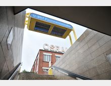 Blick von einer U-Bahn-Treppenaufgang mit geländerten Stufen auf das blau-gelbe Schild „U-Bhf Paulsternstraße“. Im Hintergrund ist ein rotes Backsteingebäude mit dem Schriftzug „OSRAM“ und hellem Himmel zu sehen.