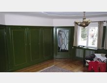 Ein traditionelles Zimmer mit grünen, holzverkleideten Einbauschränken, einem verglasten Schrank mit geometrischem Muster, hellem Holzboden und einem großen Fenster mit beigefarbenen Vorhängen. An der Decke hängt ein klassischer Kronleuchter, und im Vordergrund steht ein Tisch mit einem übergeworfenen Kleidungsstück.