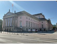 Außenansicht eines historischen Gebäudes mit klassizistischer Architektur, bekannt als Opernhaus mit rosa Fassade, Säulen, Statuen auf dem Dach und lateinischer Inschrift auf dem Giebel. Im Vordergrund sind eine Straße mit Fahrradspur und ein gepflasterter Platz zu sehen.