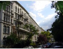 Straßenszene mit historischen Wohngebäuden im Stil des 19. Jahrhunderts, gekennzeichnet durch helle Fassaden mit dekorativen Verzierungen, Balkonen und großen Fenstern. Auf beiden Seiten der Straße stehen Bäume, während parkende Autos den Straßenrand säumen.