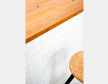 Eine Nahaufnahme zeigt eine Holztheke mit einer glatten Oberfläche auf einer Wand aus weißen rechteckigen Fliesen in Ziegelform. Im Vordergrund ist teilweise ein runder Holzsitz auf einem schwarzen Metallgestell zu sehen.