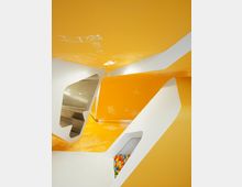 Eine moderne Innenraumgestaltung mit leuchtend gelben Wänden und weißen geometrischen Strukturen, verziert mit skizzierten Figuren und Motiven. Im Vordergrund befindet sich eine Aussparung, die eine Fläche mit bunten Plastikbällen zeigt.