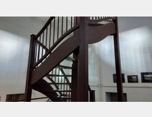 Holztreppe in einem Innenraum mit weiß gestrichenen Wänden. Im Hintergrund sind gerahmte Bilder an der Wand sowie eine Feuerlöscheinrichtung zu sehen.