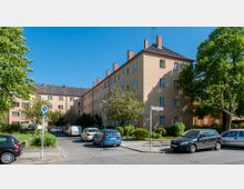 Wohnkomplex mit vierstöckigen, hell-orangefarbenen Gebäuden und dunklen Schrägdächern, umgeben von Bäumen und Sträuchern. Im Vordergrund eine ruhige Straße mit parkenden Autos und einem Straßenschild „Windsteiner Weg“.