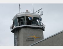 Kontrollturm des Flugplatzes Schönhagen aus grauem Beton mit einer gläsernen Aussichtsplattform. Am oberen Bereich sind Personen, Filmbeleuchtung und Kameras zu sehen.