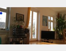 Helles Wohnzimmer mit großen Fenstern und viel Tageslicht. Der Raum enthält eine Stereoanlage mit CDs, ein schwarzes Fernsehgerät auf einem Rollgestell sowie Zimmerpflanzen und an den Wänden hängende Bilder.