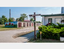 Straßenecke in einer Wohngegend mit einem Wegweiser „Kiefernweg“, im Vordergrund eine Hecke und ein Hauseingang, im Hintergrund eine Reihe von Garagen mit bemaltem Mohnblumen-Motiv und ein hoher Industrieschornstein.