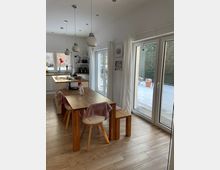 Heller, moderner Küchen- und Essbereich mit Holztisch, Stühlen und Hocker, einer Kücheninsel sowie mehreren hängenden Pendelleuchten. Große Fenster und eine Schiebetür mit weißen Vorhängen öffnen den Blick auf eine verschneite Terrasse und den Garten.