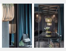 Ein modernes, stilvolles Interieur mit einem langen Esstisch aus dunklem Holz, darüber zwei Hängeleuchten mit geriffeltem Design. Im Hintergrund sind blaue Vorhänge, eine Wand mit geometrischen Mustern sowie ein gemütlicher Sitzbereich mit Polstern sichtbar.