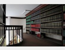 Ein Innenraum einer Bibliothek mit hohen Holzregalen, die mit Büchern in verschiedenen Farben (schwarz, rot, grün) gefüllt sind. Im Vordergrund befindet sich ein Geländer aus Holz mit vertikalen Streben, das eine Treppe begrenzt.