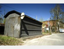 Eine einfache Metallgarage mit Flachdach und rostigen Stellen, daneben ein altes Backsteingebäude mit Fenstern und überwachsenem Efeu. Im Hintergrund befinden sich Bäume und ein klarer blauer Himmel.