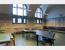 Ein historischer Seminarraum mit hohen Rundbogenfenstern, verzierten Wandpaneelen mit Blumenmustern und mehreren Tischen und Stühlen. Die Fenster lassen viel Tageslicht herein, während moderne Hängelampen den Raum zusätzlich beleuchten.