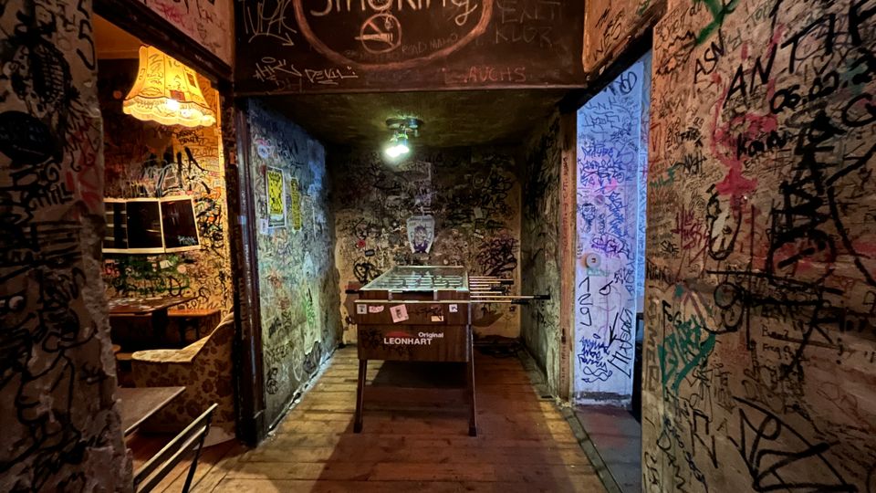 Schmaler Innenraum eines alternativen Bars oder Clubflurs mit Holzboden, dessen Wände und Decke vollständig mit Graffiti und Schriftzügen bedeckt sind. In der Mitte steht ein Tischkicker unter einer Deckenlampe; links hängt eine Stehlampe, oben ist ein „No Smoking“-Hinweis auf die Decke gemalt.