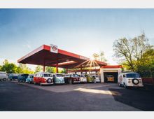 Eine Tankstelle mit einem roten Dach im Retro-Stil, unter dem mehrere bunte, klassische Volkswagen-Bullis geparkt sind. Im Hintergrund sind grüne Bäume und ein klarer blauer Himmel mit Sonnenschein zu sehen.