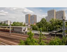 Bahnhofsumgebung mit Bahngleisen im Vordergrund, einer S-Bahn auf dem Bahnsteig und einer Treppe rechts. Im Hintergrund stehen mehrere hohe Wohngebäude mit orangenen und grauen Fassaden, umgeben von Bäumen und Grünflächen.