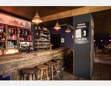 Rustikale Bar mit Holztresen und Holzbarhockern, dekoriert mit Hängelampen im Industriestil. Im Hintergrund befinden sich Regale mit Gläsern und Flaschen, eine Wand mit violettem Anstrich sowie Sitzbereich mit Tischen und Stühlen.