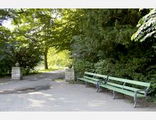 Grüne Parkbänke stehen auf einem gepflasterten Weg, der von dichten Bäumen und Sträuchern gesäumt ist. Im Hintergrund sind zwei steinerne Säulen mit Kugeldekorationen sowie weitere Bäume sichtbar, die Schatten spenden.