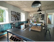 Industrieküche mit Betonwänden und -decke, großen Hängelampen und einer zentralen Arbeitsfläche mit schwarzer Metalloberfläche. Die Wände sind teils mit blauen Fliesen verkleidet, und Fenster lassen Tageslicht einfallen. Einfache Dekoration und Küchengeräte verleihen der Szene einen funktionalen Charakter.