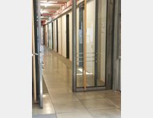 Ein moderner Büroflur mit Glastüren, weißen Wänden und einer hellen Bodenfliese. An der Decke verlaufen sichtbare rote Rohrleitungen, und links sind Türen und Informationsaushänge erkennbar.