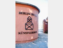 Runder, rosa-ockerfarbener Gebäudeteil mit der Aufschrift "Berlin Buch Künstlerhof" und einem modernen Kunstsymbol aus Metall an der Wand; im Hintergrund ein gepflasterter Weg und ein roter Ziegelsteinbogen.