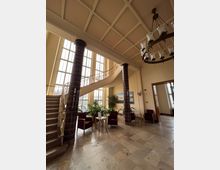 Großzügiger Eingangsbereich mit Marmorböden, hohen Fenstern und einer geschwungenen Treppe. Zwei dunkle Säulen aus glänzenden Kacheln flankieren den Raum, der mit mehreren Sitzgruppen, Pflanzen und einer großen Hängelampe ausgestattet ist.