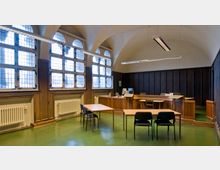 Innenansicht eines historischen Raumes mit hölzernem Richtertisch, einfachen Tischen und Stühlen sowie hohen Bleiglasfenstern. Die gewölbte Decke und Holzvertäfelung an den Wänden unterstreichen den traditionellen Charakter.