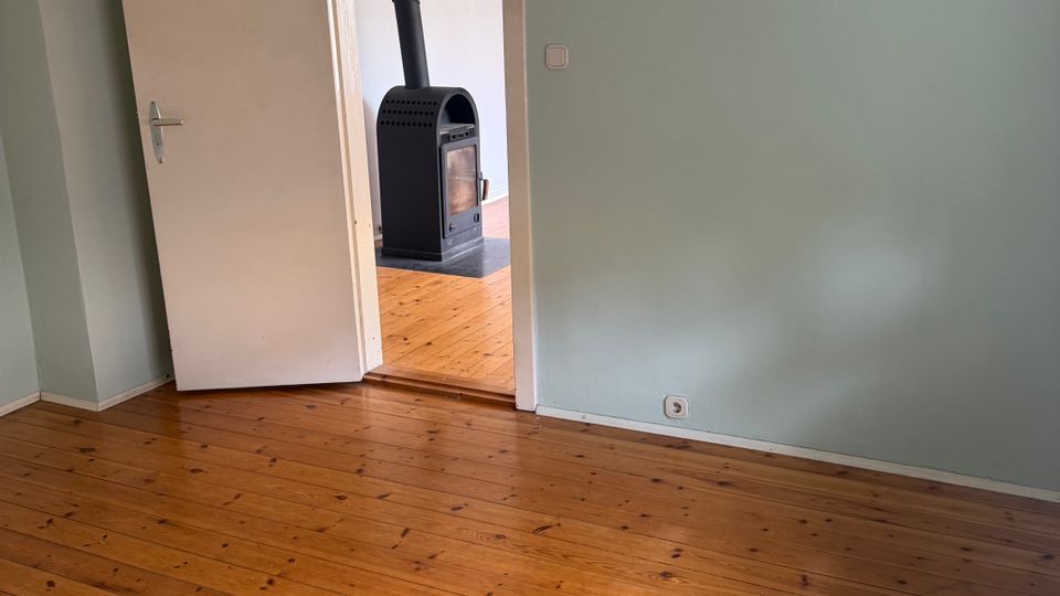 Leerer Innenraum mit hellgrün gestrichenen Wänden und breiten Holzdielen, neben einer offenen Tür. Durch den Türrahmen ist ein angrenzender Raum mit einem schwarzen Kaminofen und Ofenrohr zu sehen.