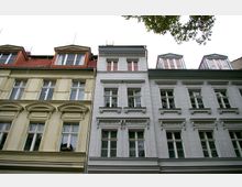 Fassadenansicht mehrerer historischer Altbau-Wohnhäuser mit verzierten Fensterrahmen aus Stuck, hellen Fassaden und Dachgauben mit roten oder grauen Dächern. Im Vordergrund ragen Äste eines Baumes in das Bild.