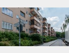 Mehrstöckiges Wohngebäude mit hellbeiger Fassade, zahlreichen rechteckigen Fenstern und kleinen Balkonen, die mit roten Kanten eingefasst sind. Im Vordergrund befindet sich ein Straßenschild mit der Aufschrift „Immenweg“, eine Fußgängerzone mit Sträuchern und Bäumen ist zu sehen, daneben verläuft eine schmale Straße mit geparkten Autos.