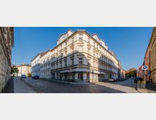 Eckgebäude in einer städtischen Umgebung mit heller Fassade und mehreren Stockwerken im historischen Baustil, umgeben von gepflasterten Straßen und parkenden Autos. Eine Seitenstraße führt links entlang, rechts ist eine weitere Straße mit Wohnhäusern zu sehen, während blauer Himmel den Hintergrund bildet.