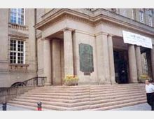 Die Abbildung zeigt den Eingang eines historischen Gebäudes mit einer breiten Treppe und mehreren Säulen. Direkt neben der Tür ist eine große Gedenktafel an der Fassade angebracht, und im Hintergrund sind rechteckige Fenster zu sehen.