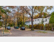 Wohnstraße in einem Wohngebiet mit herbstlich gefärbten Bäumen, parkenden Autos und zweistöckigen weißen Häusern mit roten Ziegeldächern. Auf dem Gehweg und der Straße liegen herabgefallene Blätter, Straßenschilder markieren die Kreuzung von „Devriendtweg“ und „Marienstraße“.