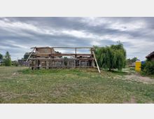 Holzstruktur mit offenem Rahmen, teils von Planen bedeckt, stehend auf einer grasbewachsenen Fläche. Rechts im Hintergrund ein gelbes, mobiles Toilettenhäuschen und ein Baum mit hängenden Ästen, unter teilweise bewölktem Himmel.