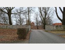 Einfahrt zu einem Park oder Friedhof mit asphaltierter Zufahrt zwischen niedrigen Steinmauern und einem geöffneten Metalltor. Dahinter stehen hohe, unbelaubte Bäume; im Hintergrund sind rote Ziegeldächer und ein Ziegelgebäude zu sehen.