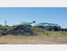 Eine Biogasanlage mit großen, grünen kuppelförmigen Behältern und silbernen Silos im Hintergrund, umgeben von Vegetation und einem Kieshaufen im Vordergrund. Der Himmel ist klar und blau.