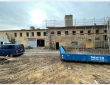 Großes, zweistöckiges Gebäude mit gelber Backsteinfassade, teilweise eingerüstet für Renovierungsarbeiten. Im Vordergrund ein blauer Transporter und ein großer blauer Abfallcontainer auf einem sandigen Bauplatz.