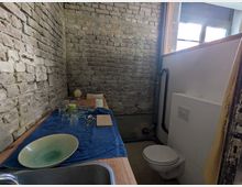 Kleiner Raum mit unverputzter Ziegelwand, einer Spüle und Arbeitsplatte links, auf der Geschirr auf einem blauen Handtuch trocknet. Rechts befindet sich eine Toilette vor einer halbhohen Trennwand, daneben ein Fenster mit Tageslicht.