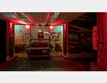 Industrieartiger Innenraum mit düsterer Beleuchtung in Rot- und Grüntönen, dekoriert mit Wandmalereien, darunter ein männliches Figurenmotiv mit Krone. Im Vordergrund steht ein massives Metallmöbelstück, während im Hintergrund bunte Wandstreifen und Poster erkennbar sind.