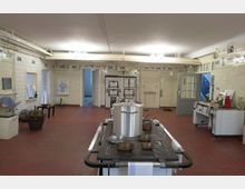 Große historische Küche mit cremefarben gefliesten Wänden, roten Bodenfliesen und sichtbaren Rohren an der Decke. In der Mitte steht ein großer Herd mit Töpfen und Pfannen, während an den Wänden alte Küchengeräte und Möbel wie Spülen und Regale zu sehen sind.